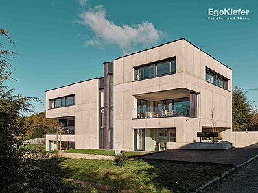 Ein modernes, kubisches Mehrfamilienhaus mit flachem Dach, großen Fenstern und Balkonen, umgeben von einer üppigen grünen Landschaft.